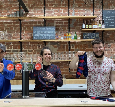 L’image montre trois personnes souriantes derrière un bar avec un fond en briques apparentes. Ils tiennent et présentent des chaussettes colorées avec des motifs. L’ambiance semble chaleureuse et conviviale, évoquant un lieu accueillant où se mêlent artisanat, commerce local et bonne humeur.