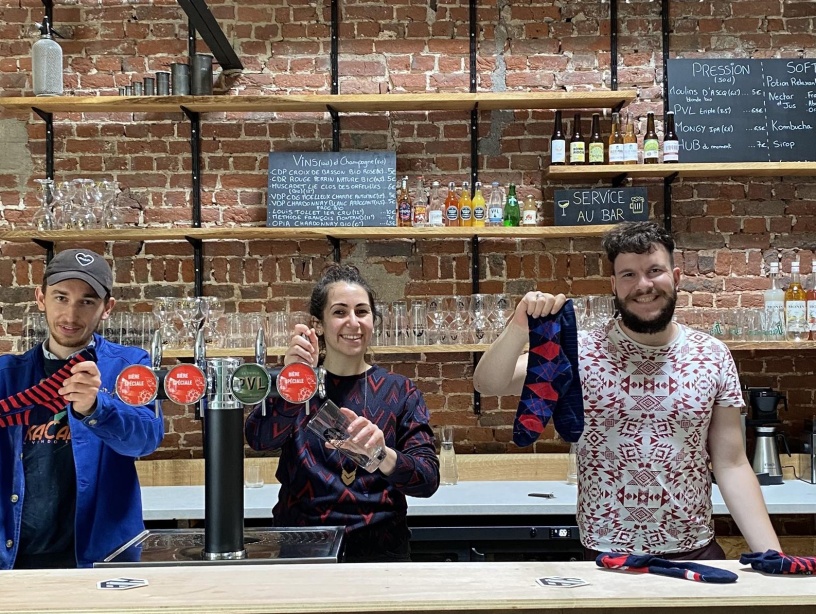 L’image montre trois personnes souriantes derrière un bar avec un fond en briques apparentes. Ils tiennent et présentent des chaussettes colorées avec des motifs. L’ambiance semble chaleureuse et conviviale, évoquant un lieu accueillant où se mêlent artisanat, commerce local et bonne humeur.